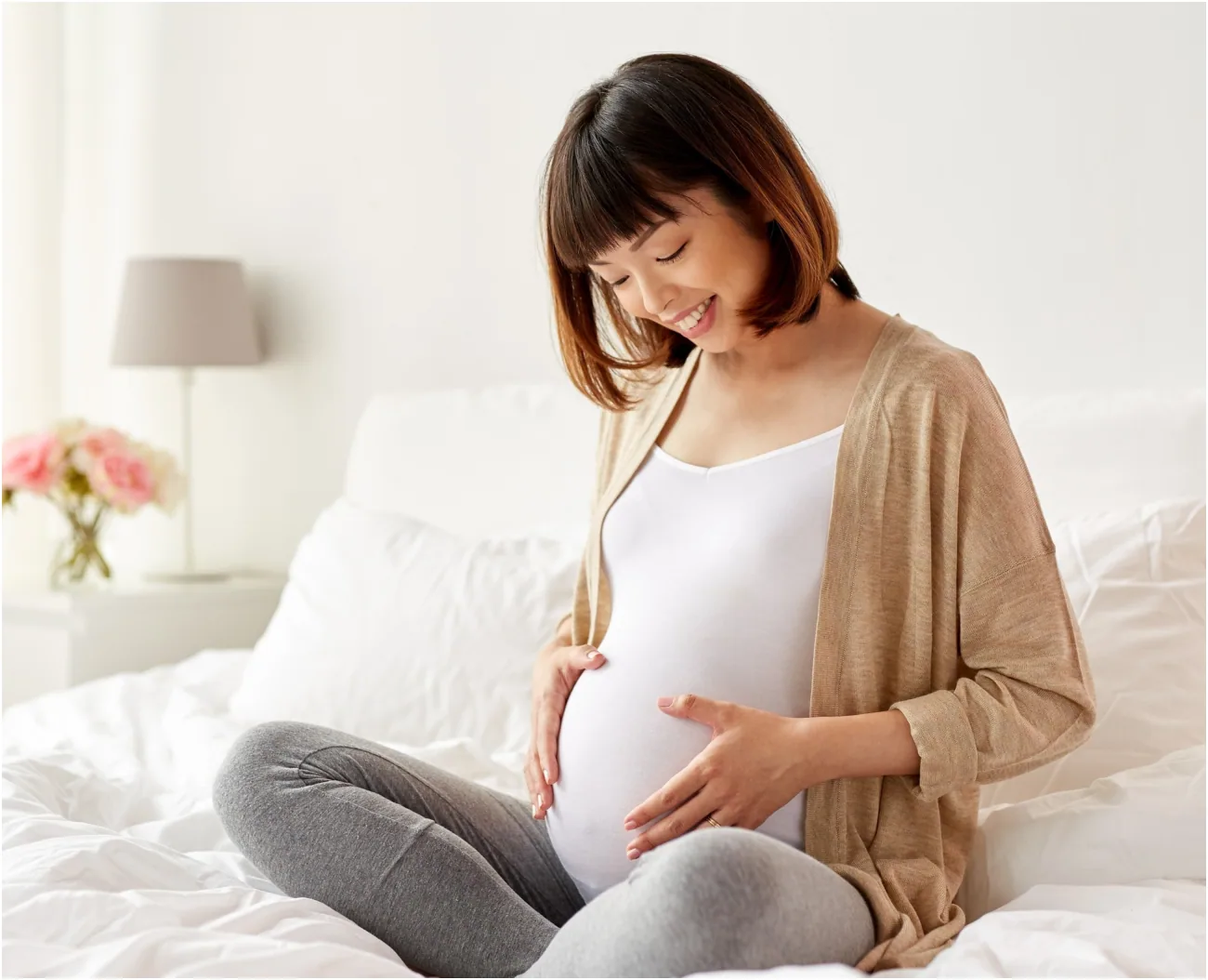 A smiling pregnant woman sitting on a bed with her hands on her belly